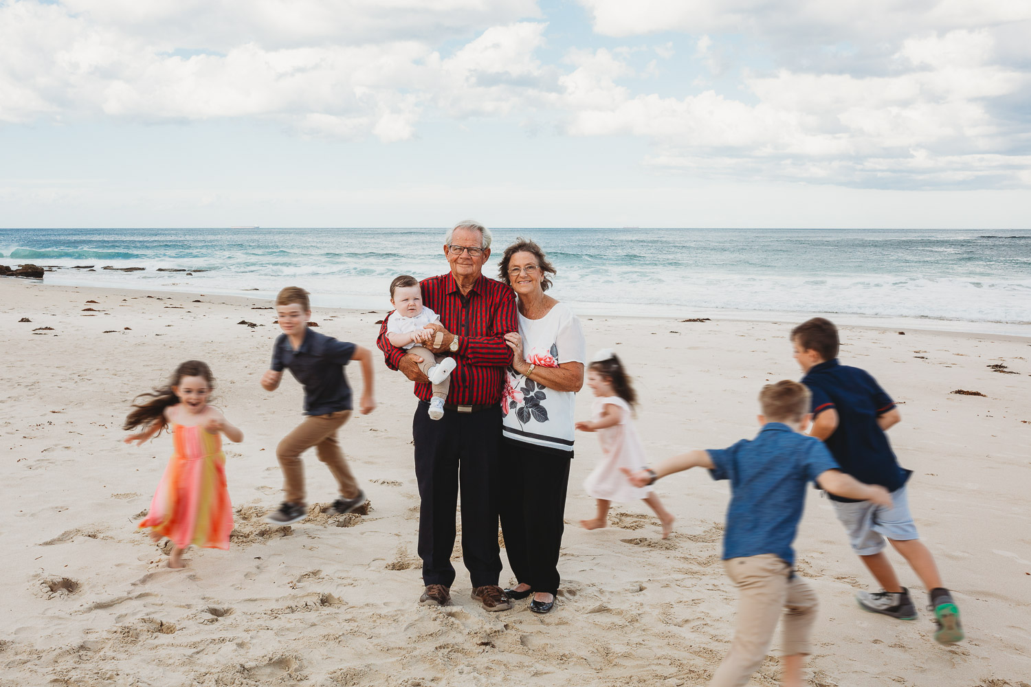 Grandparents in the middle in focus holding baby while grandkids are blurred running around them