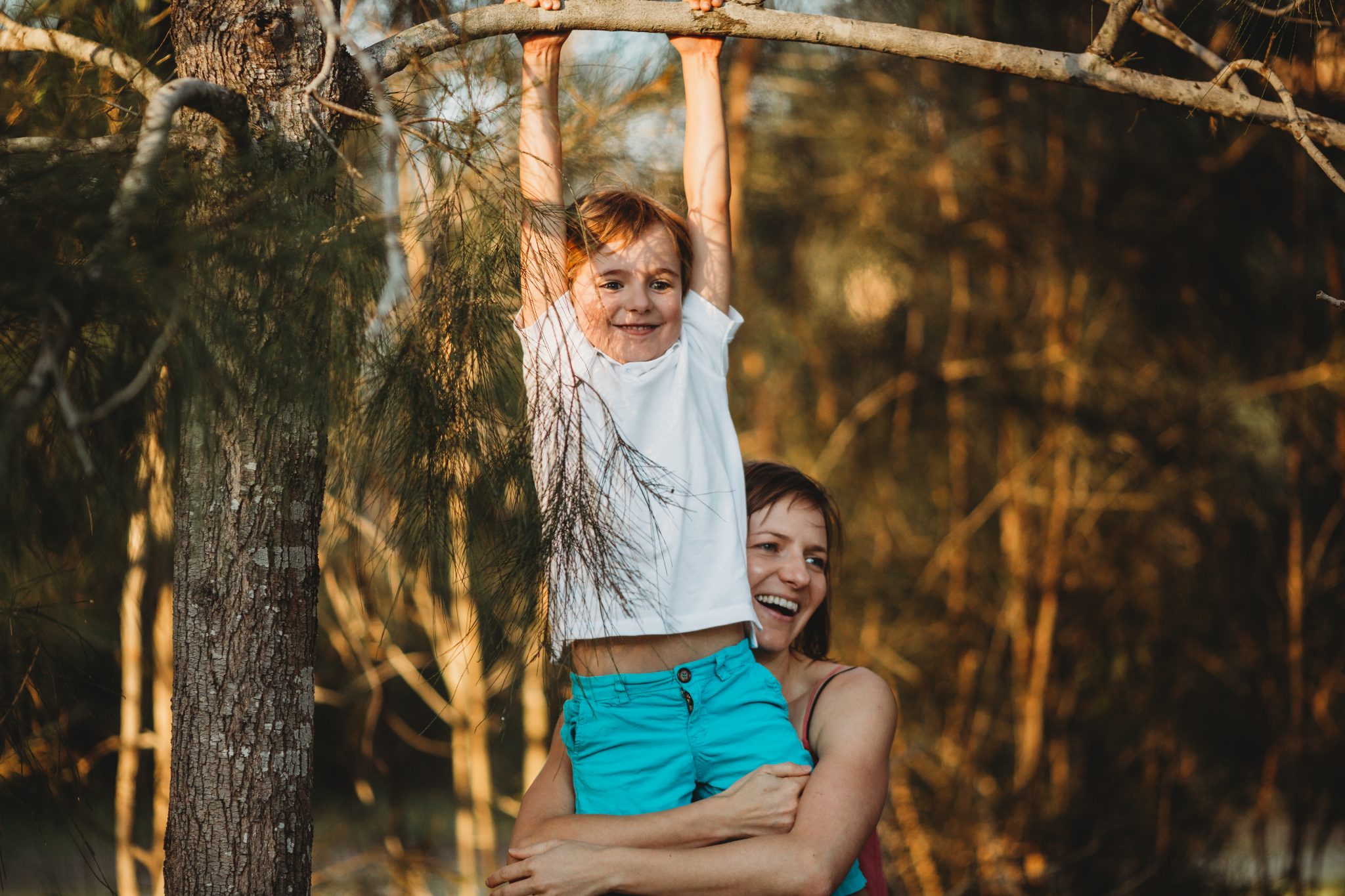 Young boy smiling, while hanging from branch as his mother supports him