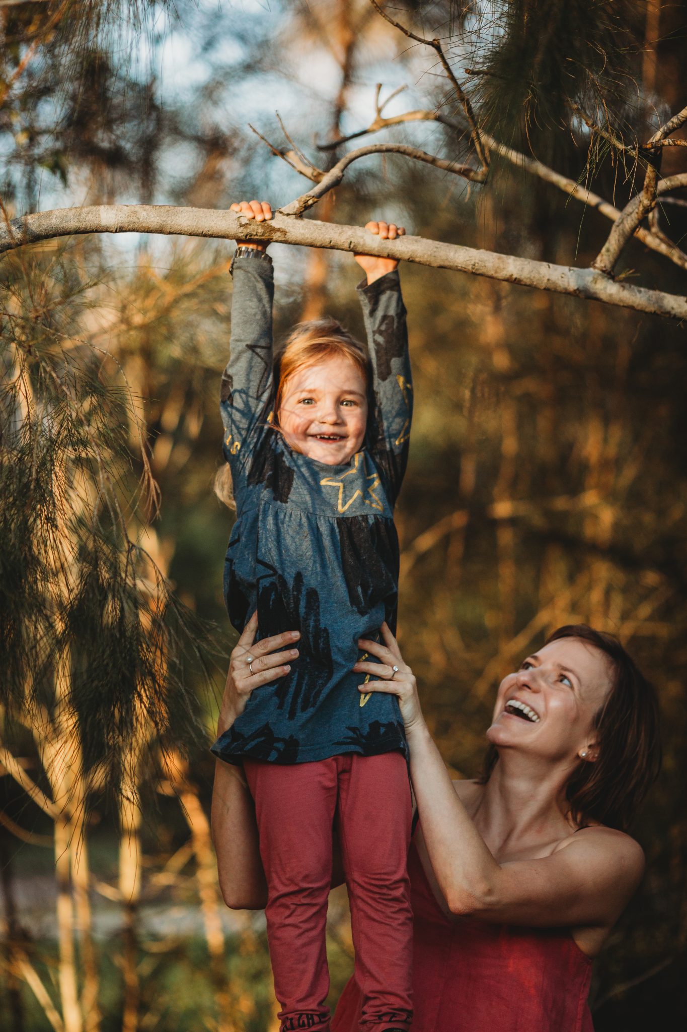 Young girl smiling, while hanging from branch as her mother supports her