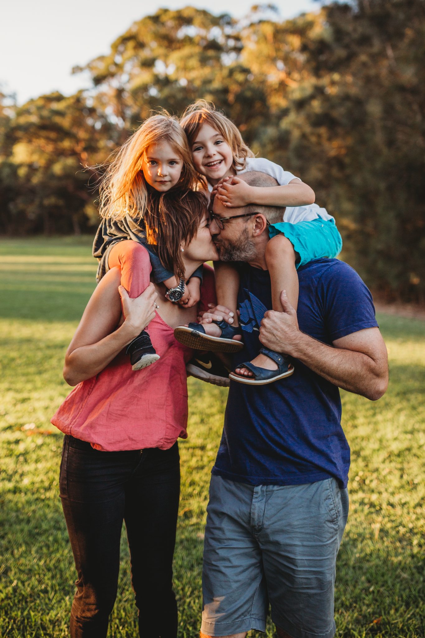 Man and woman kissing while their daughter and son sit on their shoulders, smiling at camera