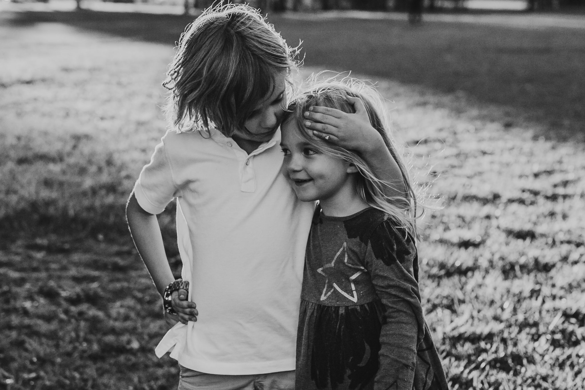 Black and white image of young boy hugging his sister side on while looking at her as she smiles