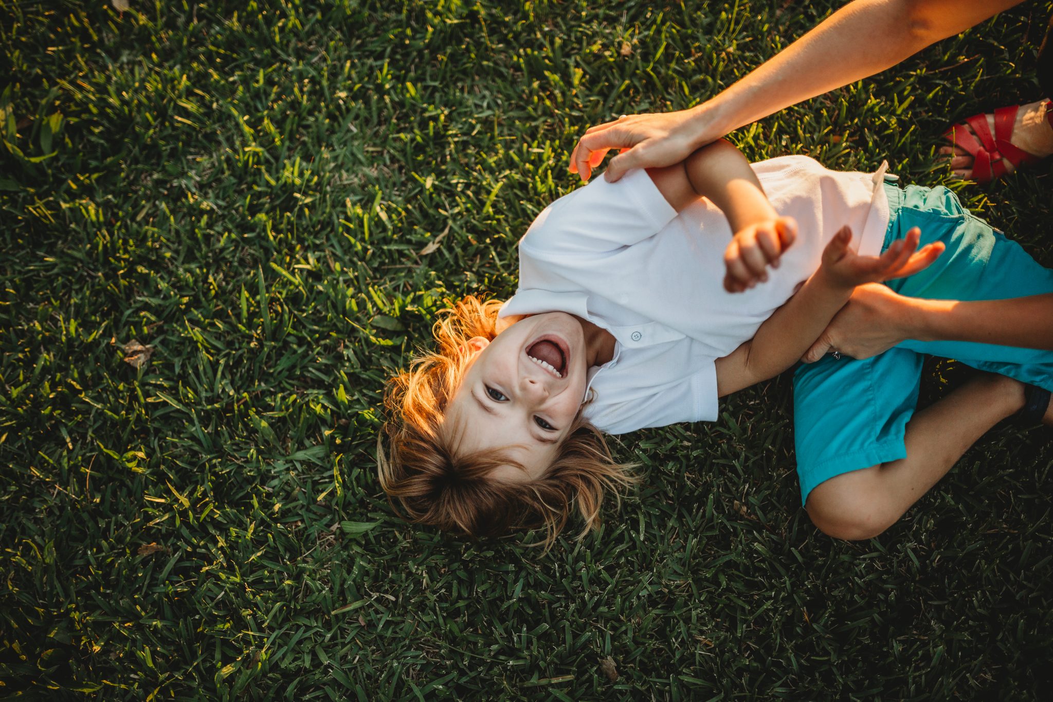 Young boy laughing, while laying on the grass, looking at the camera while being tickled by her mum