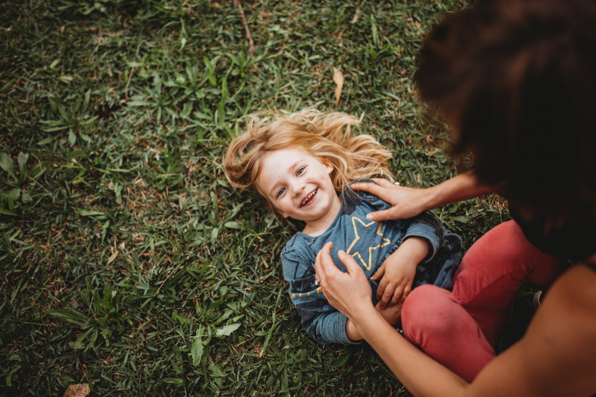 Young girl smiling, while laying on the grass, looking at the camera while being tickled by her mum
