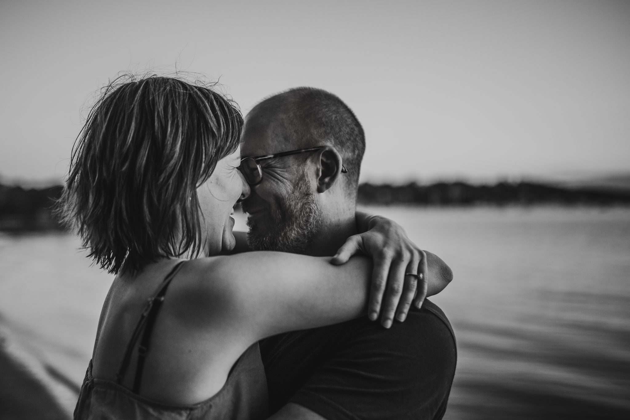 Black and white image of woman and man hugging by Lake Macquarie