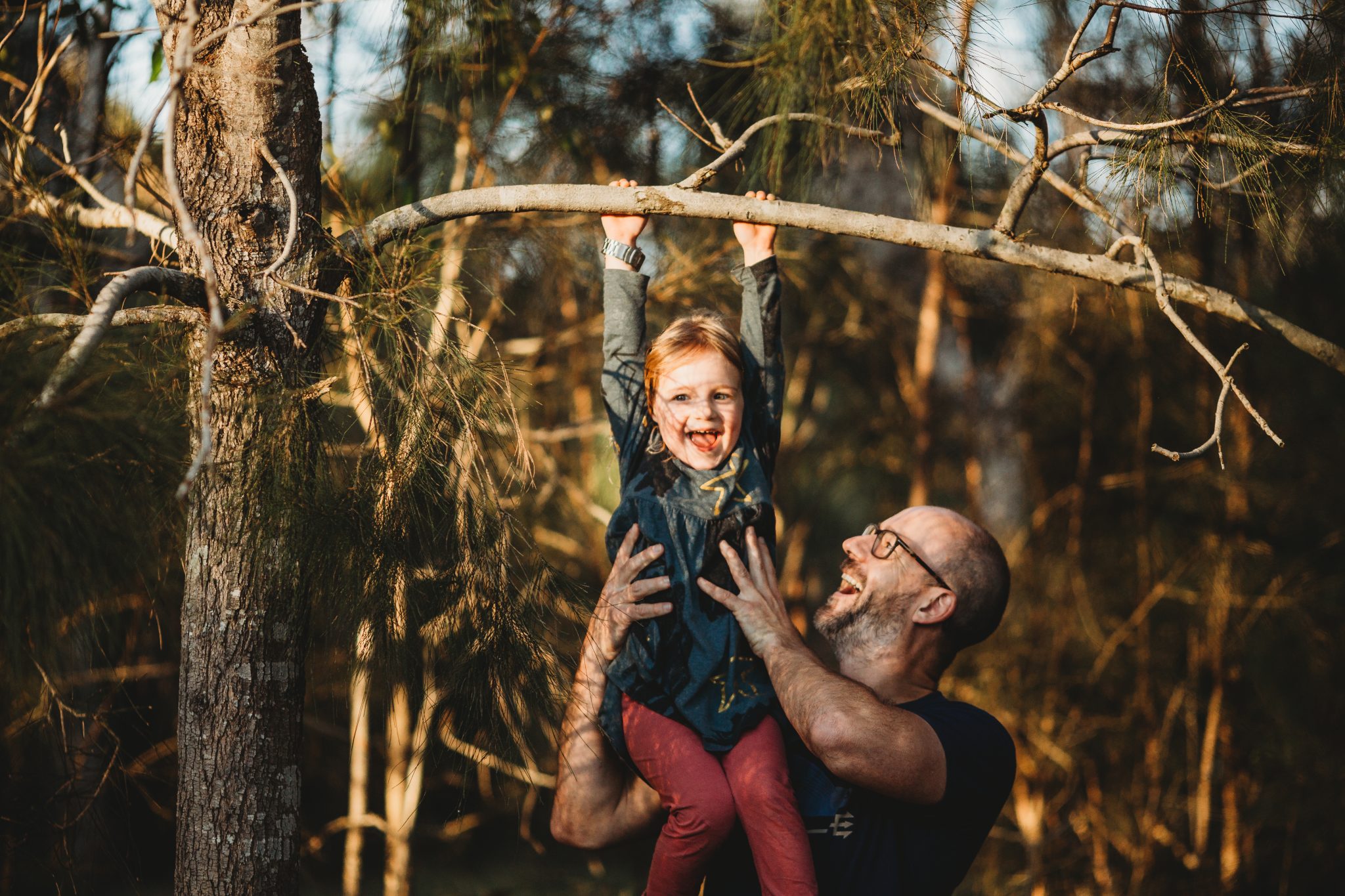 Young girl smiling while hanging off tree branch while her father holds her up