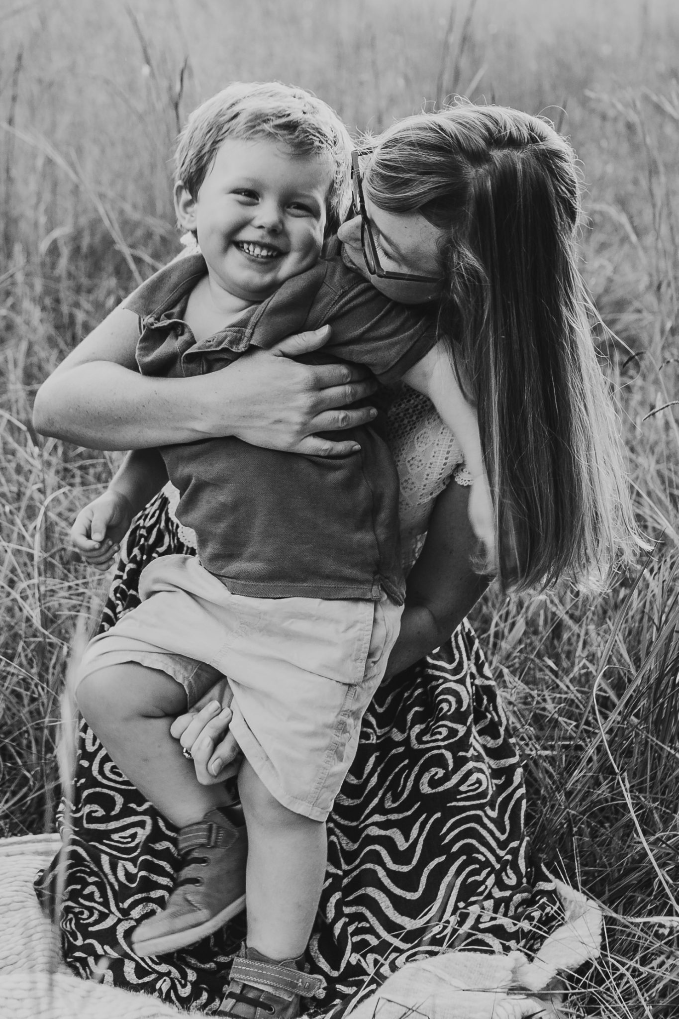 Black and white image of boy smiling as his mother hugs and kisses him