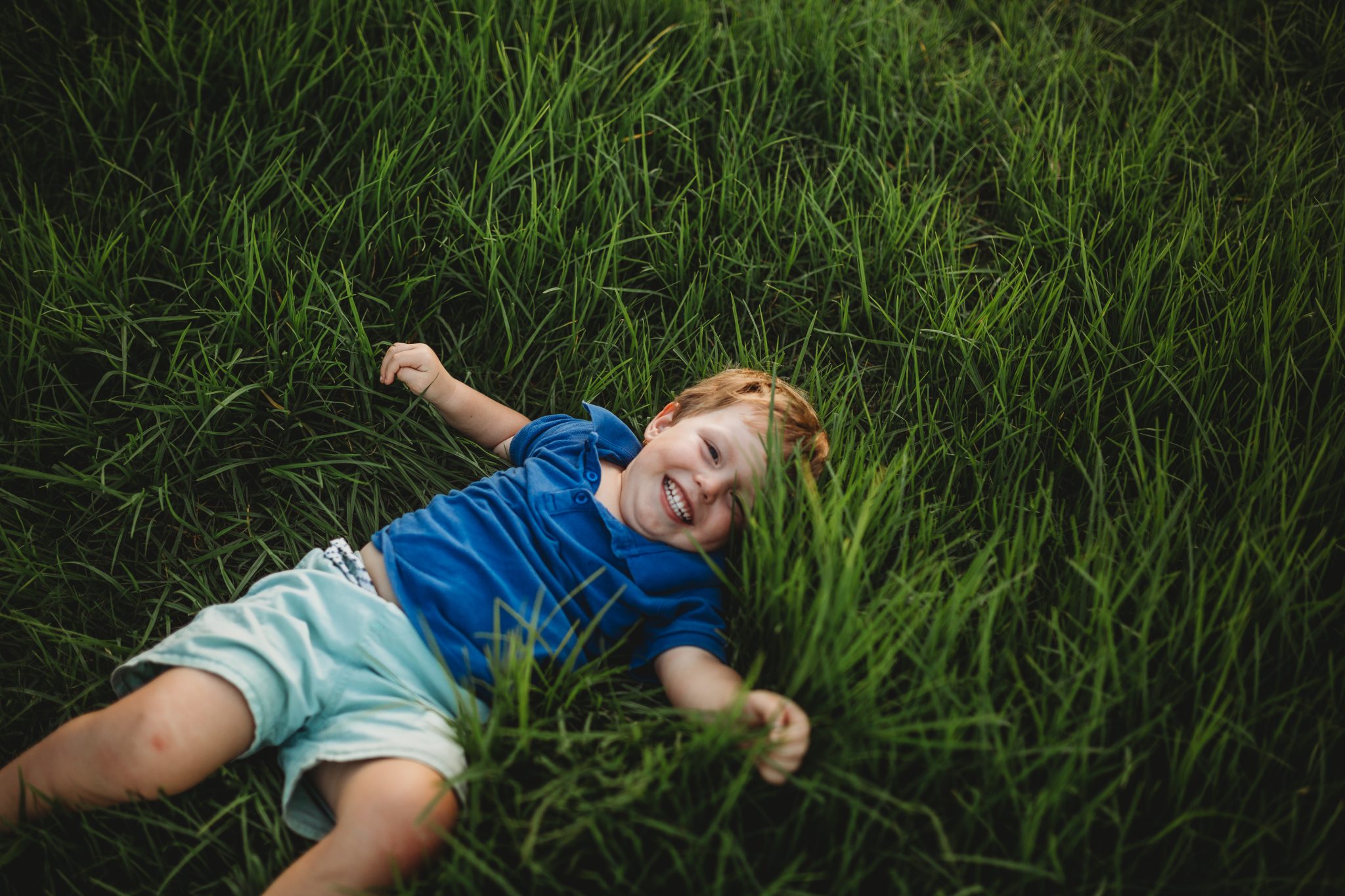 Young boy in blue shirt, laying in green grass and smiling