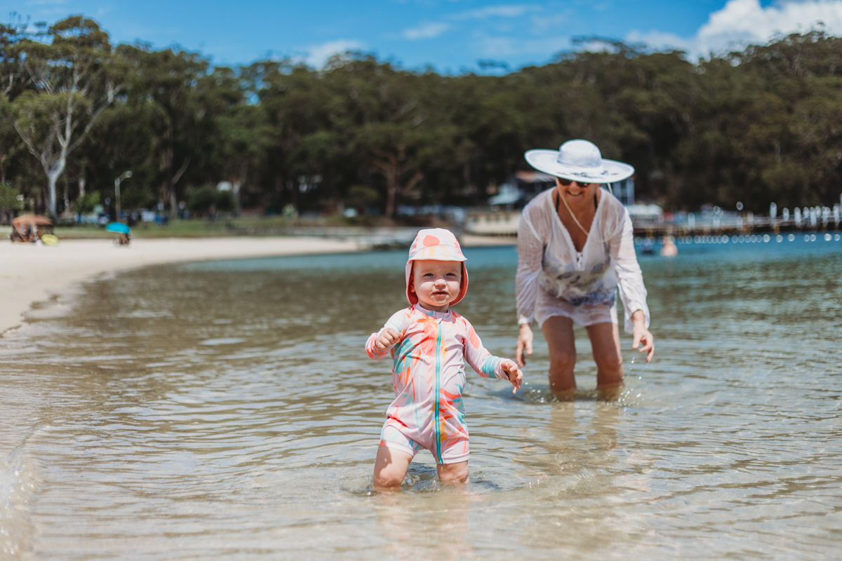 Toddler standing in water at the beach, with grandmother smiling behind her