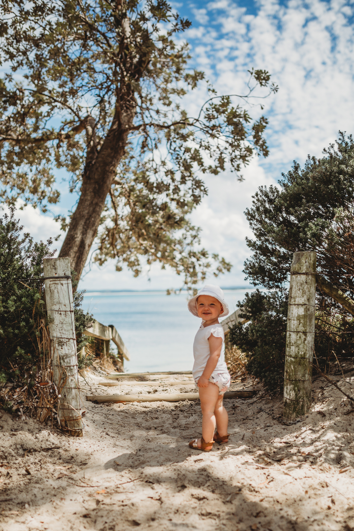 Little girl with white hat standing at pathway to beach, smiling