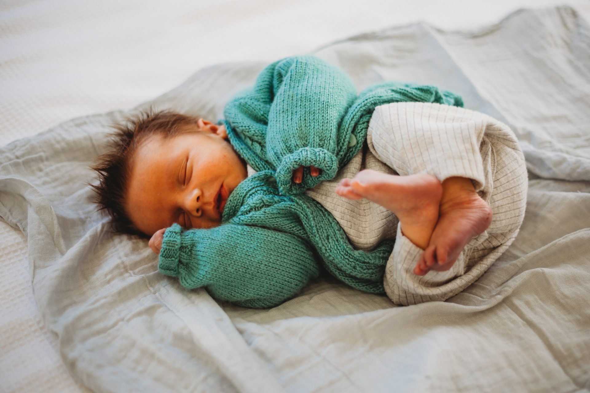 Sleeping newborn baby boy in green cardigan, laying on a grey wrap on a bed