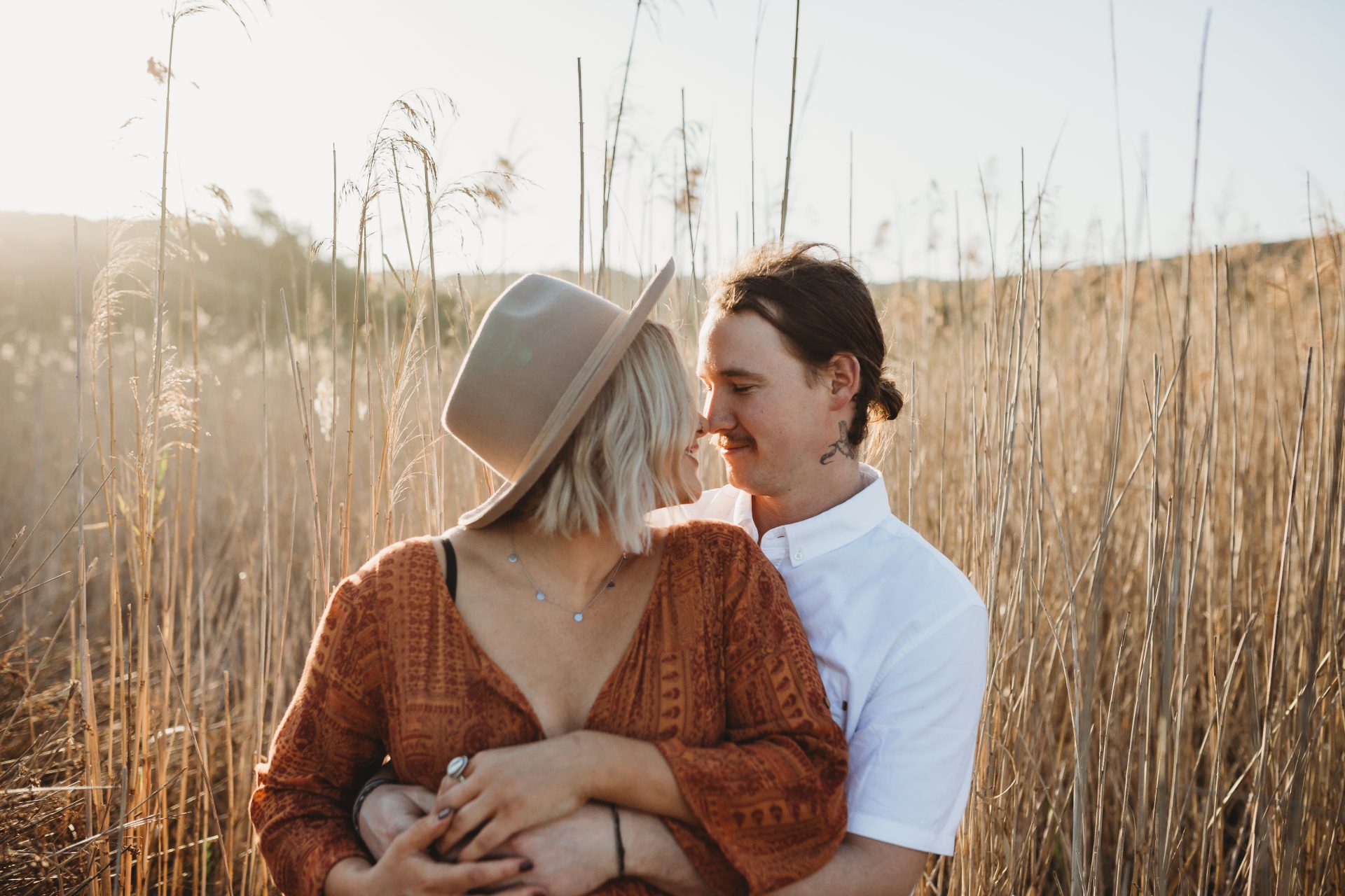 Young boho couple from Lake Macquarie touching noses, while posing in front of tall reeds at sunset