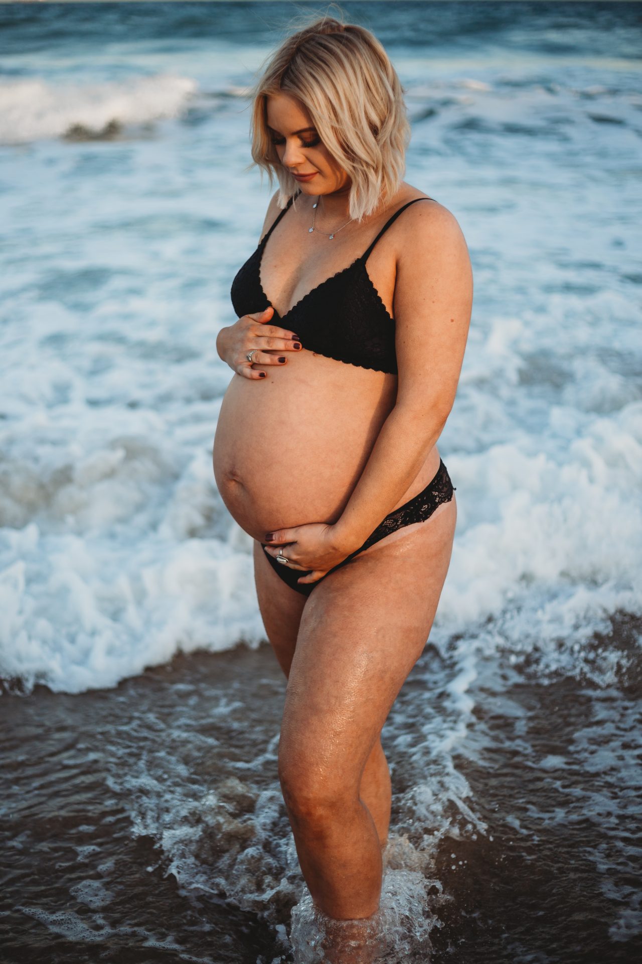 Young pregnant woman's stomach, posed in ocean at sunset, wearing black bra and underwear