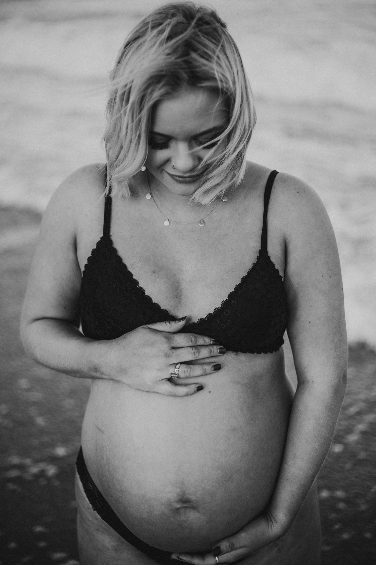 Black and white image of blonde woman looking down at her pregnant belly, while standing in the water at the beach