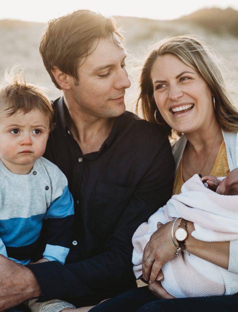 Woman laughing, holding her baby daughter, as husband looks lovingly at her while holding toddler who stares into the distance