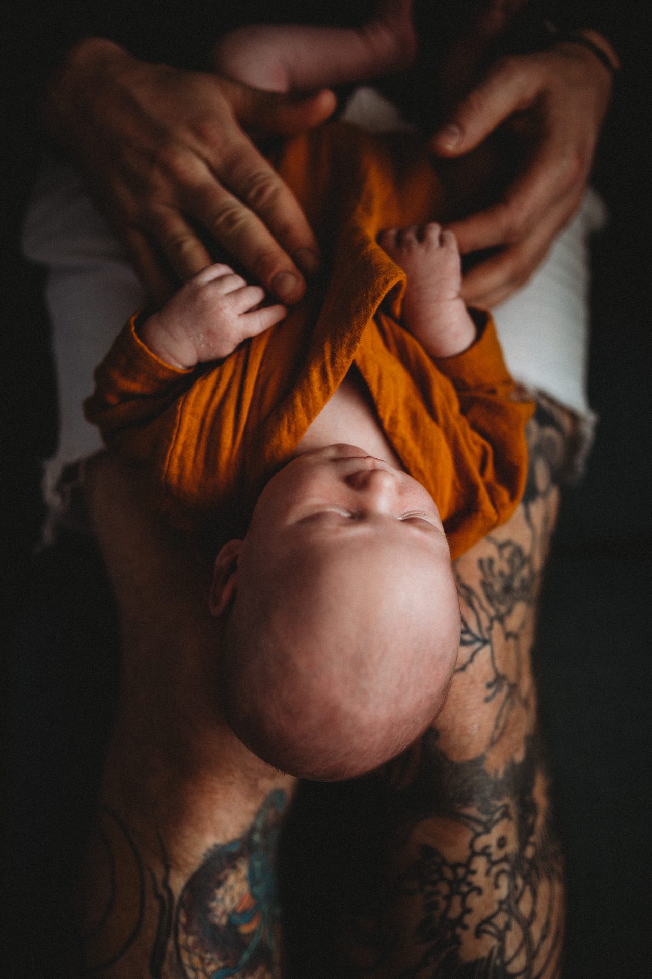 Newborn baby dressed in orange linen suit, laying on his father's tattooed legs