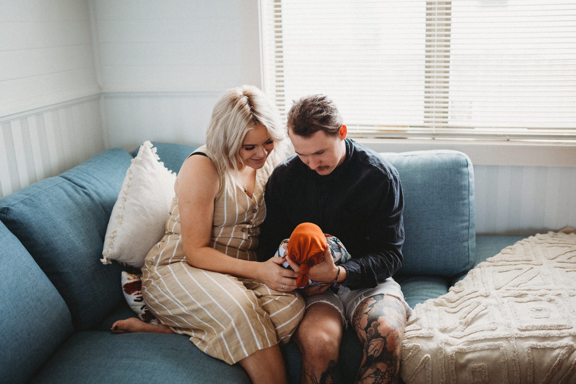 Young boho couple sitting together on couch, both holding newborn baby and looking down at him