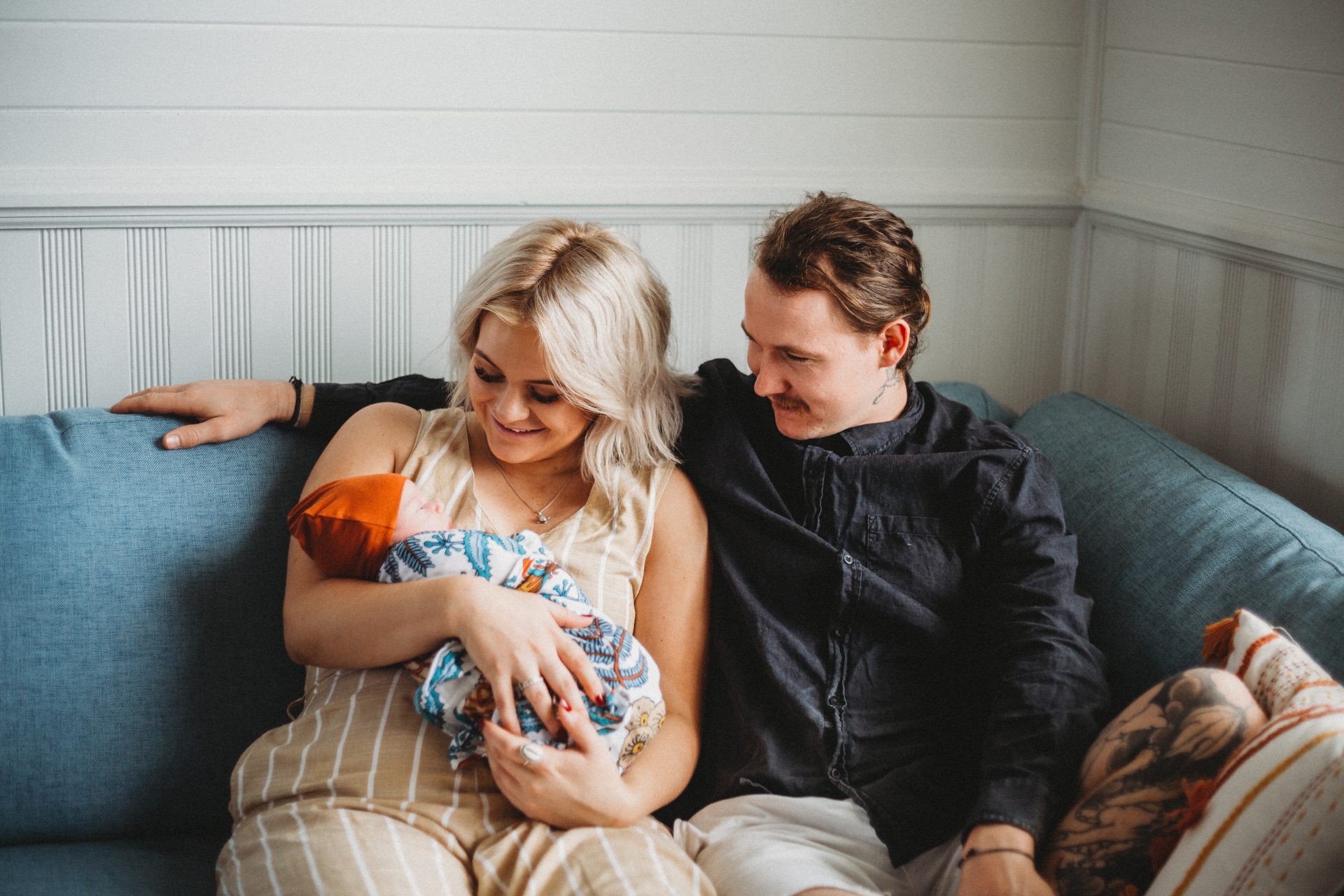 Young woman on couch holding newborn son, with her partner next to her, with his arm around her, both looking and smiling at baby