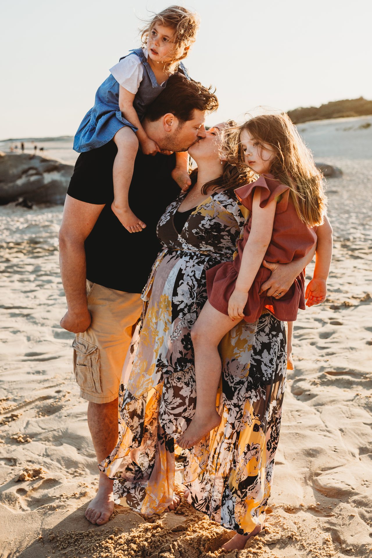 Mother and father kissing while husband has one daughter on his shoulders, and his wife is holding their other daughter on her hip