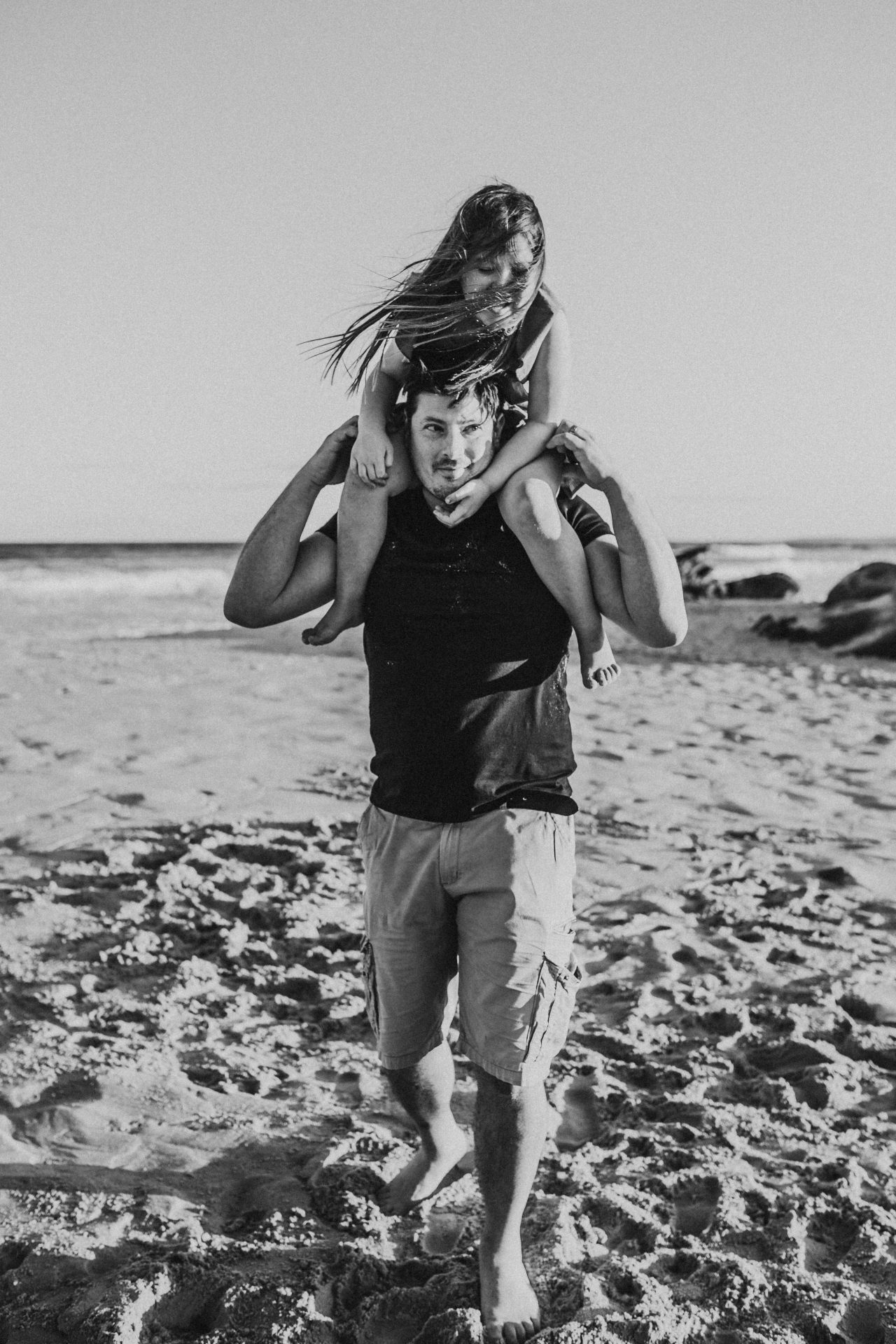 Black and white image of a young girl on her dad's shoulders, holding her hands under his chin, as he holds onto her legs