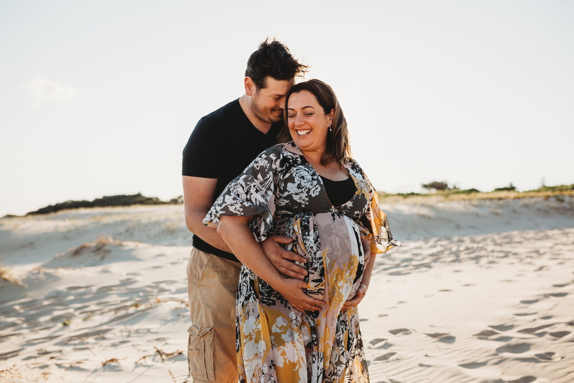 Pregnant woman standing in front of her husband smiling, as he cuddles her from behind