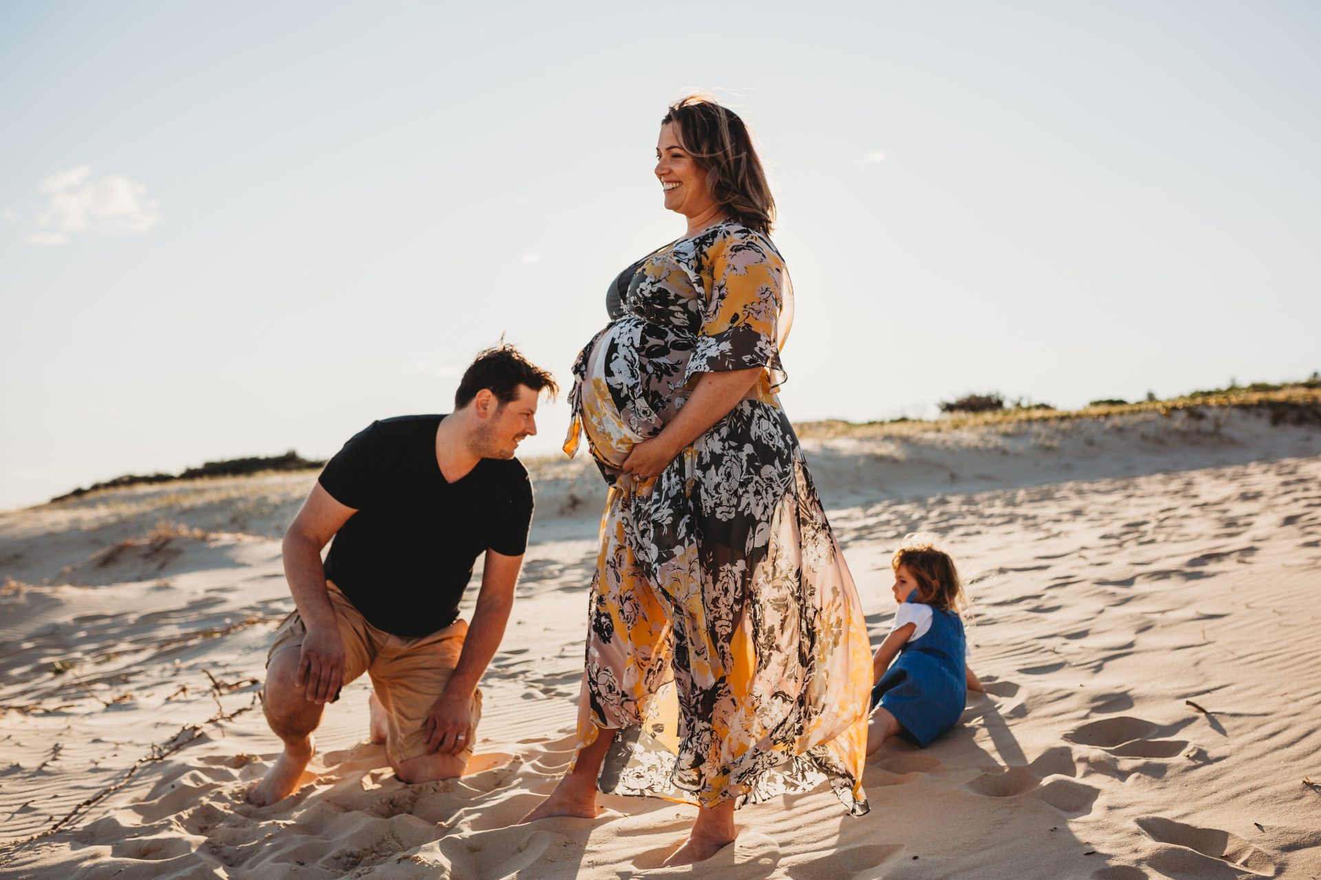 Pregnant woman at the beach, standing side-on between her husband and daughter, who are sitting on the sand