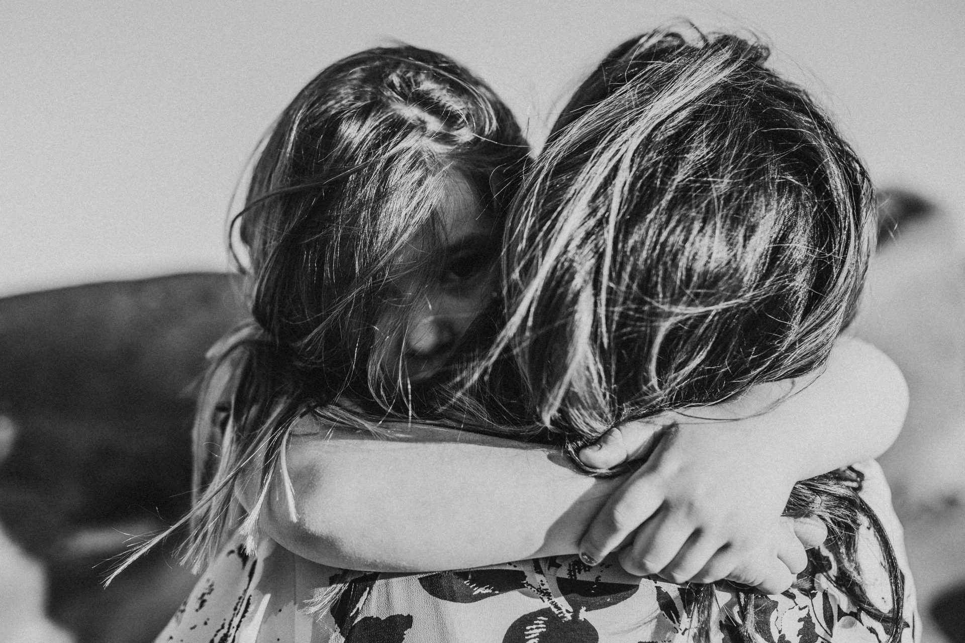 Black and white image of a little girl facing the camera as she cuddles her mother, arms around her neck
